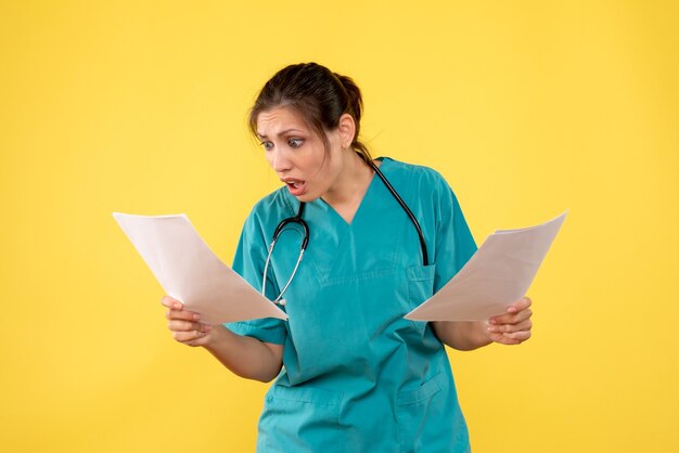 Jovem médica com camisa médica segurando uma análise de papel sobre fundo amarelo de frente