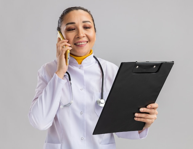 Jovem médica com jaleco branco com estetoscópio em volta do pescoço, segurando a prancheta, sorrindo enquanto fala no celular, em pé sobre uma parede branca