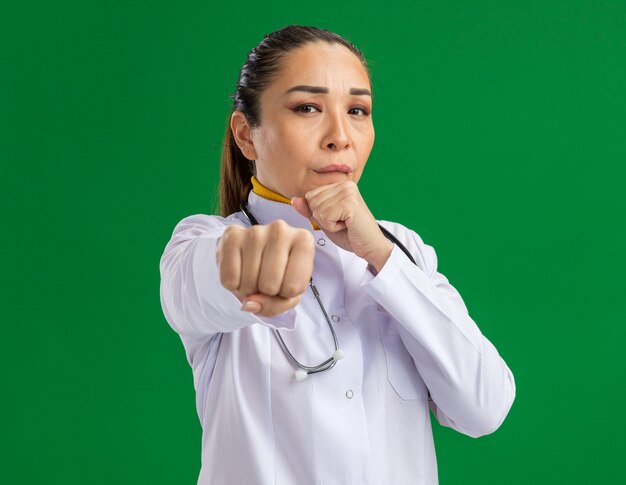 Jovem médica com jaleco branco com estetoscópio no pescoço e rosto sério, posando como um boxeador com os punhos cerrados em pé sobre a parede verde