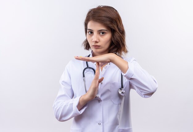 Jovem médica com jaleco branco com estetoscópio parecendo cansada fazendo gesto de pausa com as mãos