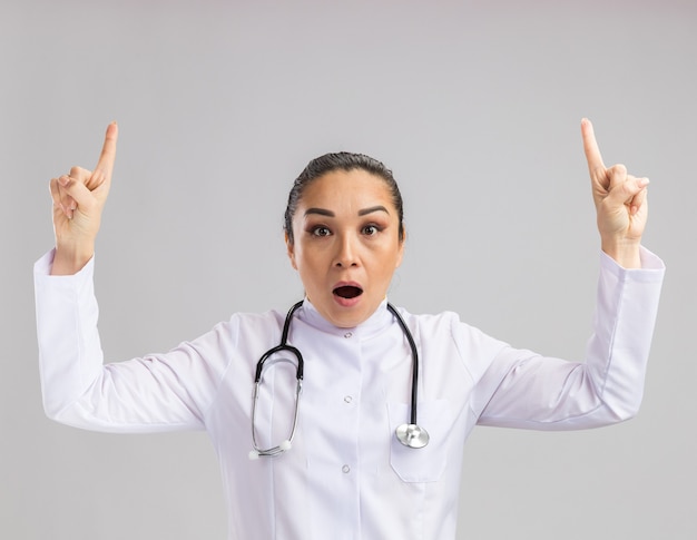 Jovem médica com jaleco branco e estetoscópio pendurado no pescoço, surpresa, mostrando os dedos indicadores em pé sobre uma parede branca