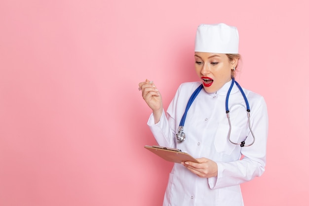 Jovem médica com terno branco e estetoscópio azul, segurando o bloco de notas no espaço rosa, vista frontal