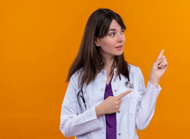 Jovem médica confiante em um manto médico com estetoscópio apontando para o lado com ambas as mãos em um fundo laranja isolado com espaço de cópia