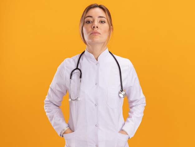 Jovem médica confiante vestindo túnica médica com estetoscópio, colocando as mãos no bolso isolado na parede amarela