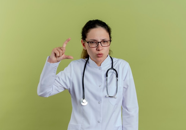 Jovem médica confiante vestindo túnica médica e estetoscópio com óculos mostrando o tamanho isolado na parede verde oliva
