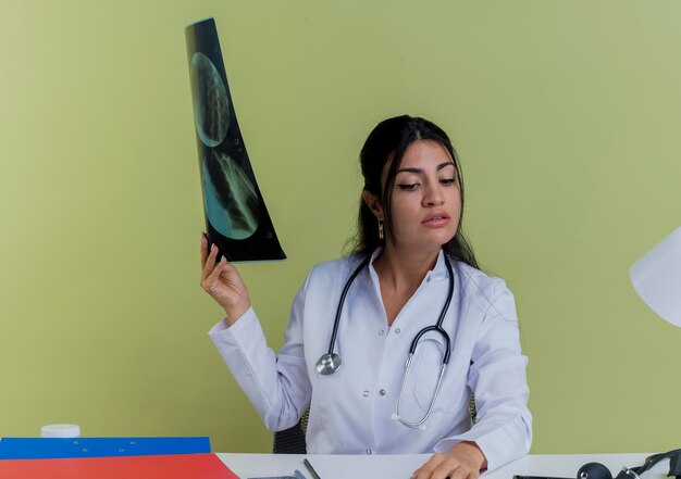 Jovem médica confiante vestindo túnica médica e estetoscópio sentada na mesa com ferramentas médicas, segurando uma foto de raio-x, colocando a mão na mesa olhando para a mesa isolada