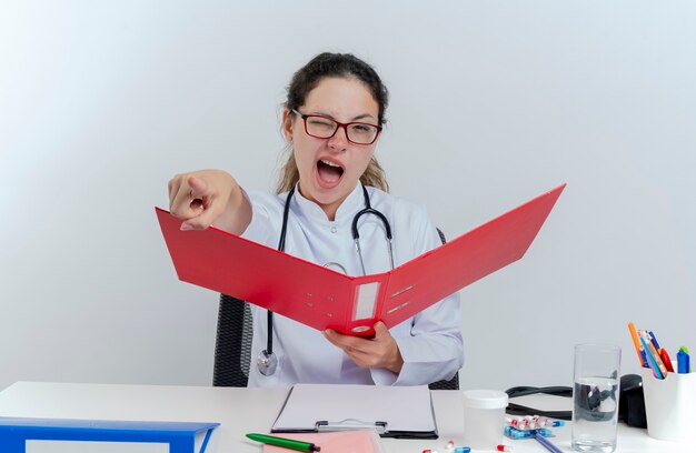 Jovem médica confiante vestindo túnica médica, estetoscópio e óculos, sentada na mesa com ferramentas médicas, segurando uma pasta, olhando e apontando piscando isolado