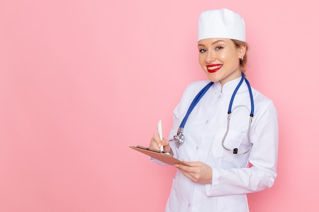 Jovem médica de terno branco com estetoscópio azul, segurando o bloco de notas com um sorriso no espaço rosa, vista frontal