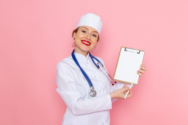 Jovem médica de terno branco com estetoscópio azul segurando o bloco de notas com um sorriso no hospital médico de medicina espacial rosa