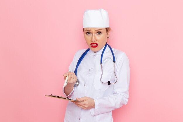 Jovem médica de terno branco com estetoscópio azul segurando o bloco de notas e olhando para a câmera no espaço rosa