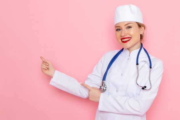 Jovem médica de terno branco com estetoscópio azul, vista frontal, posando e sorrindo no trabalho feminino do espaço rosa