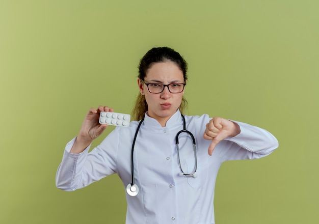 Jovem médica descontente, vestindo túnica médica e estetoscópio com óculos segurando comprimidos, mostrando o polegar para baixo isolado na parede verde oliva
