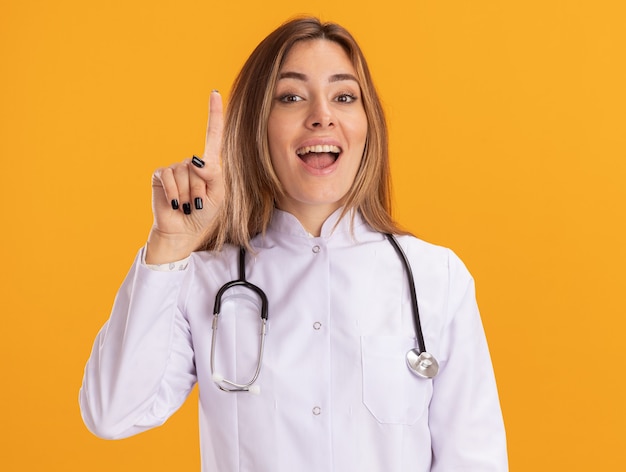 Foto grátis jovem médica impressionada usando manto médico com estetoscópio apontando para cima isolado na parede amarela