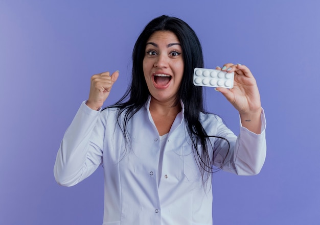 Foto grátis jovem médica impressionada usando túnica de médico, mostrando pacote de comprimidos médicos, com o punho cerrado