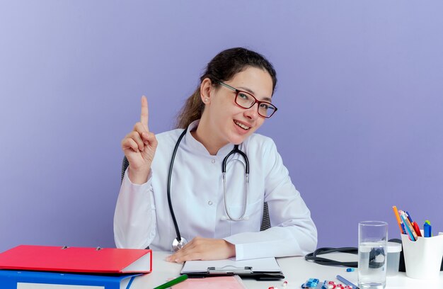 Jovem médica impressionada usando túnica médica e estetoscópio sentada à mesa com ferramentas médicas, olhando levantando o dedo isolado