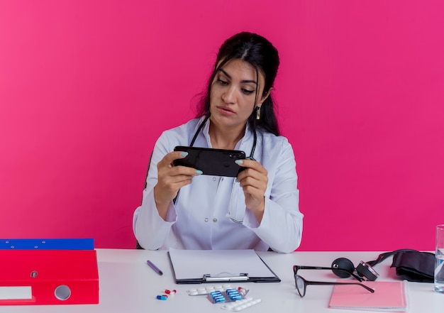 Jovem médica impressionada usando túnica médica e estetoscópio, sentada à mesa com ferramentas médicas, segurando e olhando para o telefone celular isolado na parede rosa