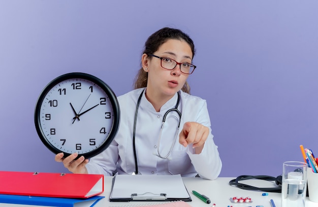 Jovem médica impressionada usando túnica médica e estetoscópio, sentada à mesa com ferramentas médicas, segurando um relógio, olhando e apontando isolado