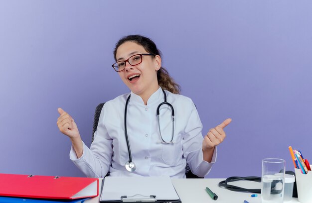 Jovem médica impressionada usando túnica médica e estetoscópio sentada na mesa com ferramentas médicas, parecendo mostrando os polegares isolados