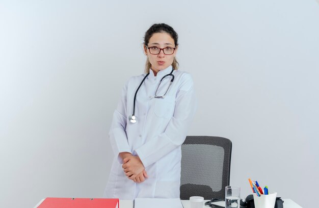 Jovem médica impressionada usando túnica médica, estetoscópio e óculos, em pé atrás da mesa com ferramentas médicas, mantendo as mãos juntas isoladas