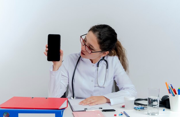Jovem médica impressionada usando túnica médica, estetoscópio e óculos, sentada na mesa com instrumentos médicos, mostrando e olhando para o telefone celular isolado
