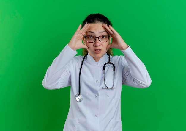 Jovem médica impressionada usando túnica médica, estetoscópio e óculos, tocando o rosto isolado na parede verde com espaço de cópia