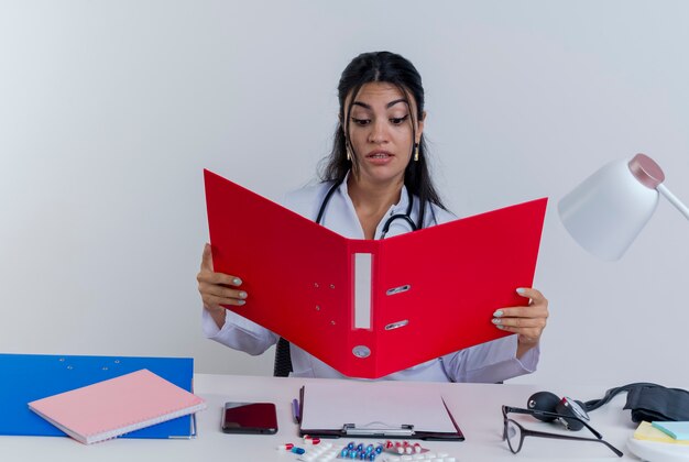 Jovem médica impressionada, vestindo bata médica e estetoscópio, sentada à mesa com ferramentas médicas, segurando e olhando para a pasta isolada