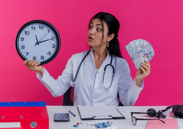 Foto grátis jovem médica impressionada vestindo túnica médica e estetoscópio sentada na mesa com ferramentas médicas segurando dinheiro e relógio olhando para o relógio isolado na parede rosa