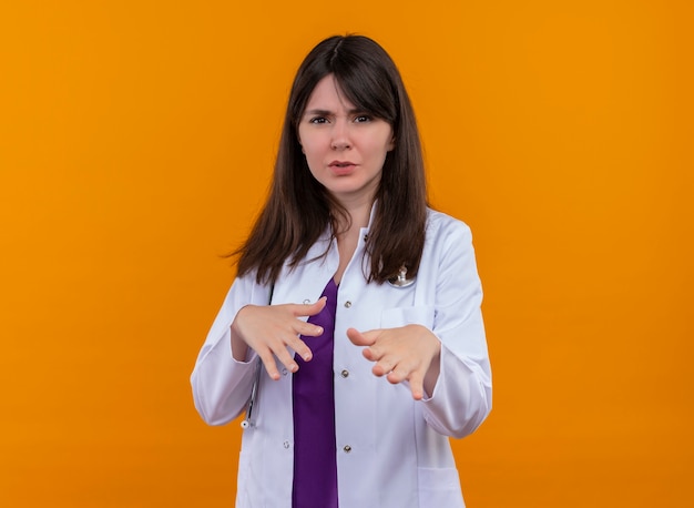 Jovem médica irritada com túnica médica com gestos de estetoscópio para longe de mim em fundo laranja isolado com espaço de cópia