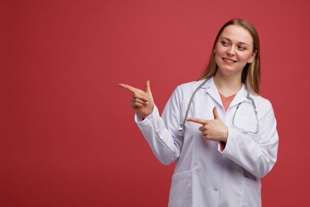 Jovem médica loira sorridente, vestindo bata médica e estetoscópio pendurado no pescoço, olhando e apontando para o lado