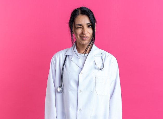Jovem médica piscando satisfeita, vestindo bata médica com estetoscópio isolado na parede rosa