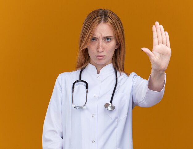 Jovem médica ruiva, carrancuda, vestindo bata médica e estetoscópio olhando para frente, fazendo gesto de parada isolado na parede laranja