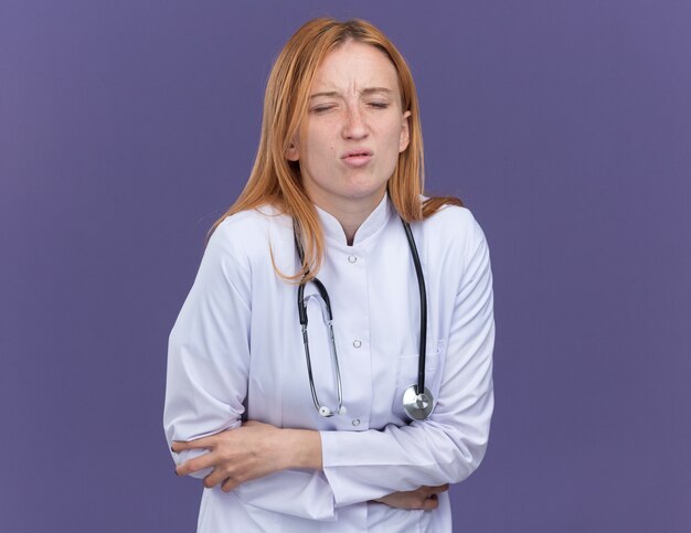 Jovem médica ruiva dolorida, vestindo túnica médica e estetoscópio, segurando a barriga com os olhos fechados, isolada na parede roxa com espaço de cópia