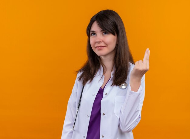 Jovem médica satisfeita com um manto médico com estetoscópio gesticula dois com os dedos em um fundo laranja isolado com espaço de cópia