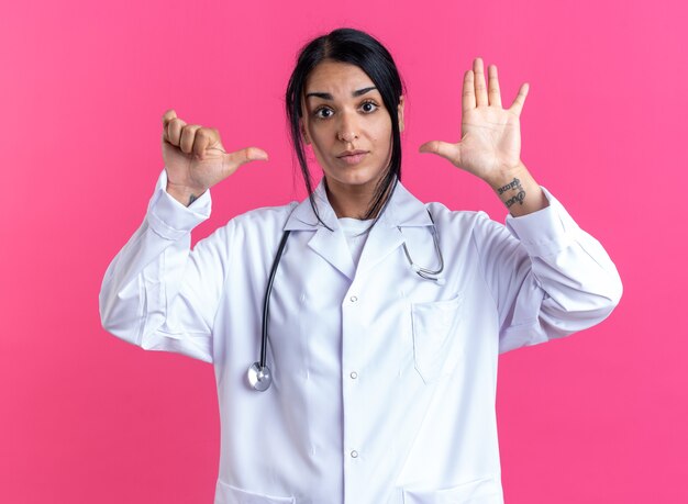 Jovem médica satisfeita usando manto médico com estetoscópio mostrando diferentes gestos isolados na parede rosa