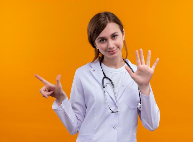 Jovem médica satisfeita usando manto médico e estetoscópio apontando para o lado esquerdo e mostrando cinco no espaço laranja isolado