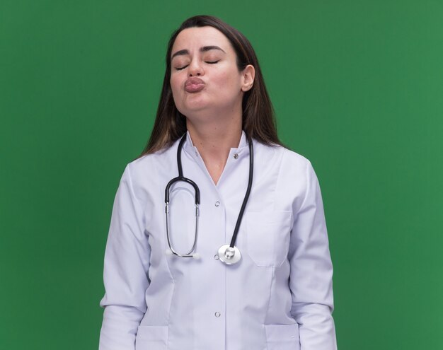 Jovem médica satisfeita vestindo túnica médica com estetoscópio e fingindo beijar no verde