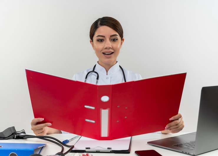 Foto grátis jovem médica satisfeita vestindo túnica médica com estetoscópio sentada na mesa de trabalho no computador com ferramentas médicas segurando e olhando para a pasta com espaço de cópia