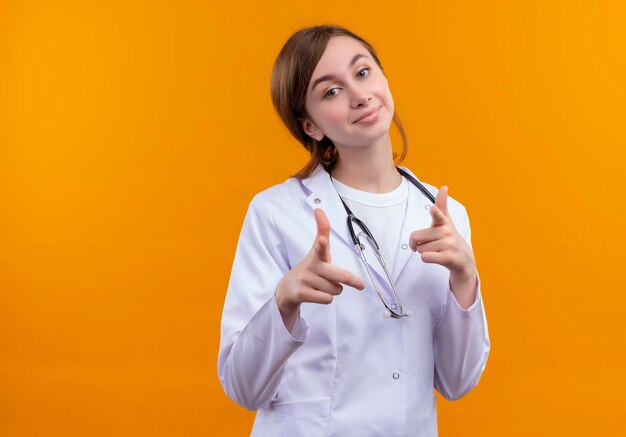 Jovem médica satisfeita vestindo túnica médica e estetoscópio fazendo seu gesto no espaço laranja isolado com espaço de cópia