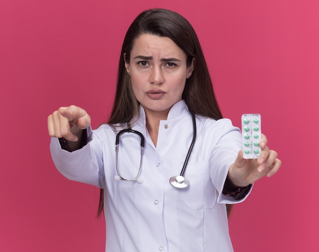 Foto grátis jovem médica séria vestindo túnica médica com estetoscópio segurando pacote de remédios e aponta para a câmera isolada na parede rosa com espaço de cópia