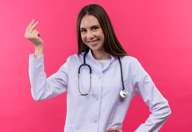 Jovem médica sorridente com bata médica de estetoscópio mostrando gesto de ponta em fundo rosa isolado