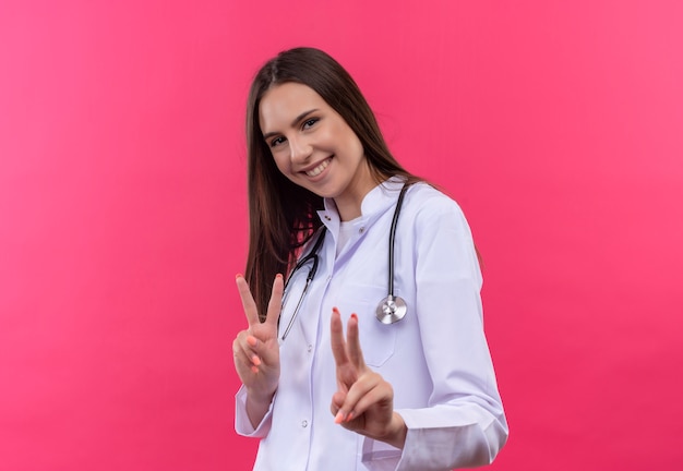 Jovem médica sorridente com bata médica de estetoscópio mostrando um gesto de paz em fundo rosa isolado