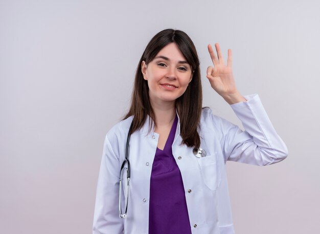 Jovem médica sorridente com manto médico com estetoscópio e gestos ok em fundo branco isolado com espaço de cópia