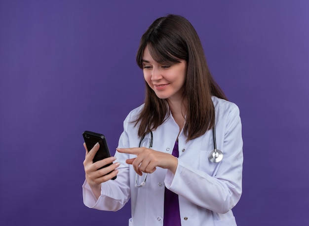 Jovem médica sorridente com manto médico com estetoscópio segura o telefone e aponta para o telefone no fundo violeta isolado com espaço de cópia