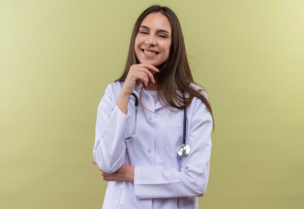 Jovem médica sorridente usando bata médica de estetoscópio e colocando a mão no queixo sobre fundo verde