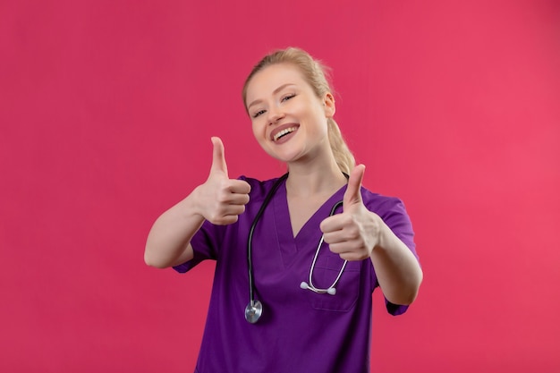 Jovem médica sorridente usando bata médica roxa e estetoscópio com os polegares para cima na parede rosa isolada