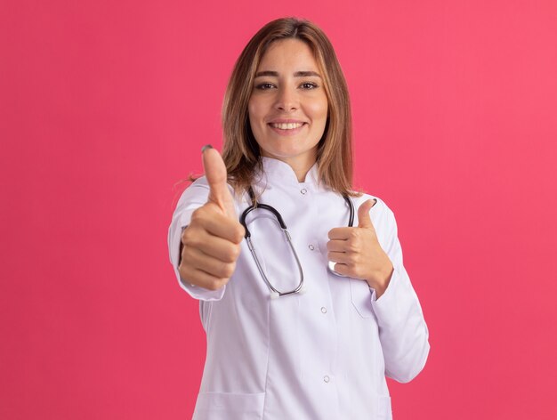 Jovem médica sorridente usando manto médico com estetoscópio mostrando os polegares isolados na parede rosa