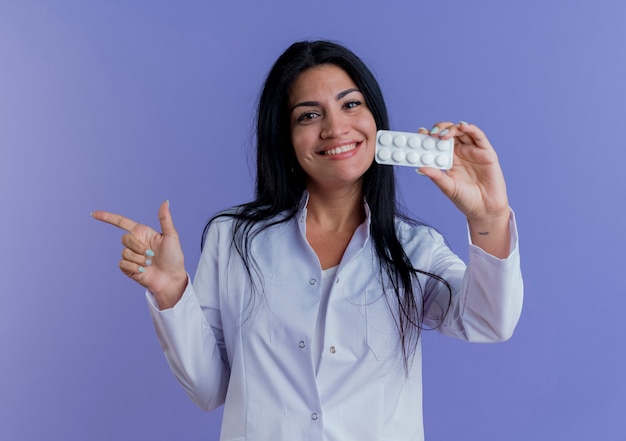 Jovem médica sorridente usando manto médico, mostrando a embalagem de comprimidos médicos, olhando apontando para o lado