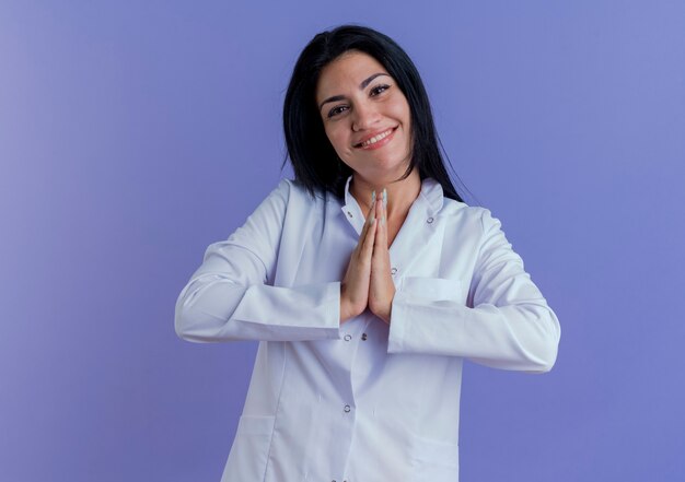 Jovem médica sorridente usando manto médico olhando para manter as mãos juntas