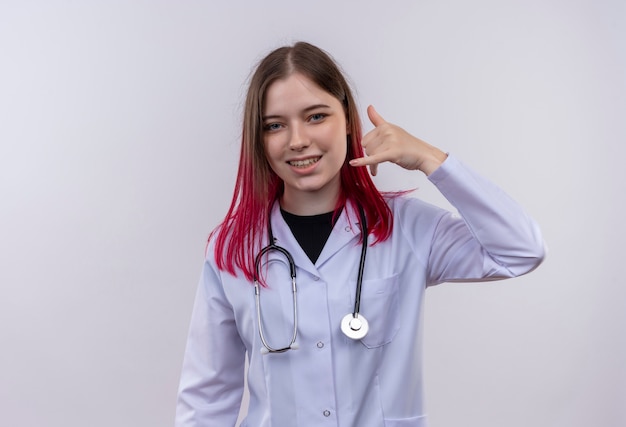 Jovem médica sorridente usando túnica médica de estetoscópio, mostrando gesto de chamada telefônica em fundo branco isolado