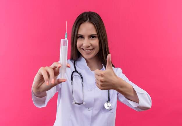 Jovem médica sorridente usando um jaleco de estetoscópio e segurando a seringa com o polegar no fundo rosa